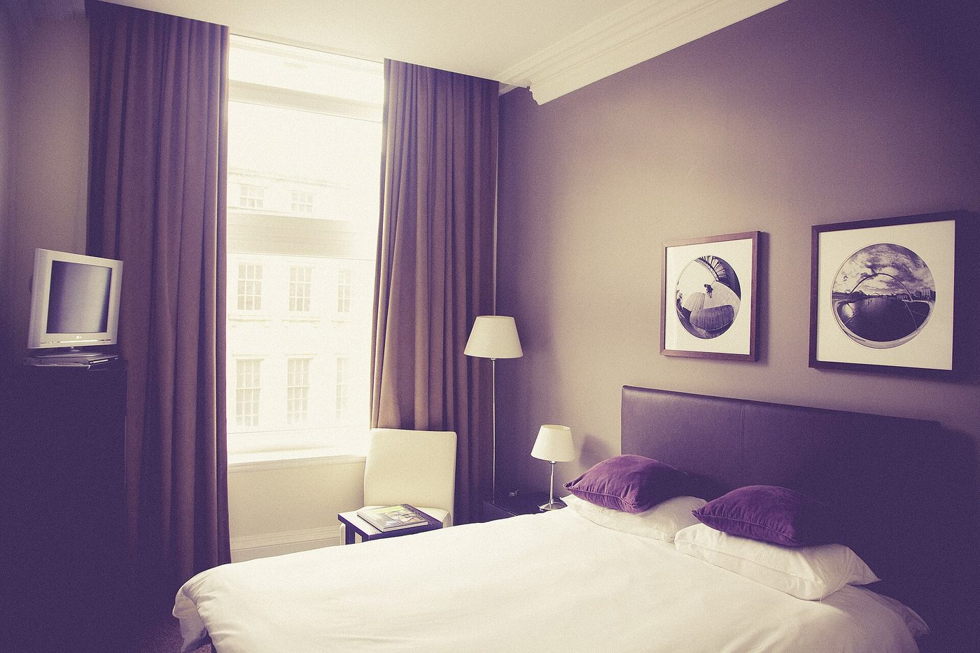 A double room with white bedding, bedside lamps, framed pictures and a tall window with long curtains.