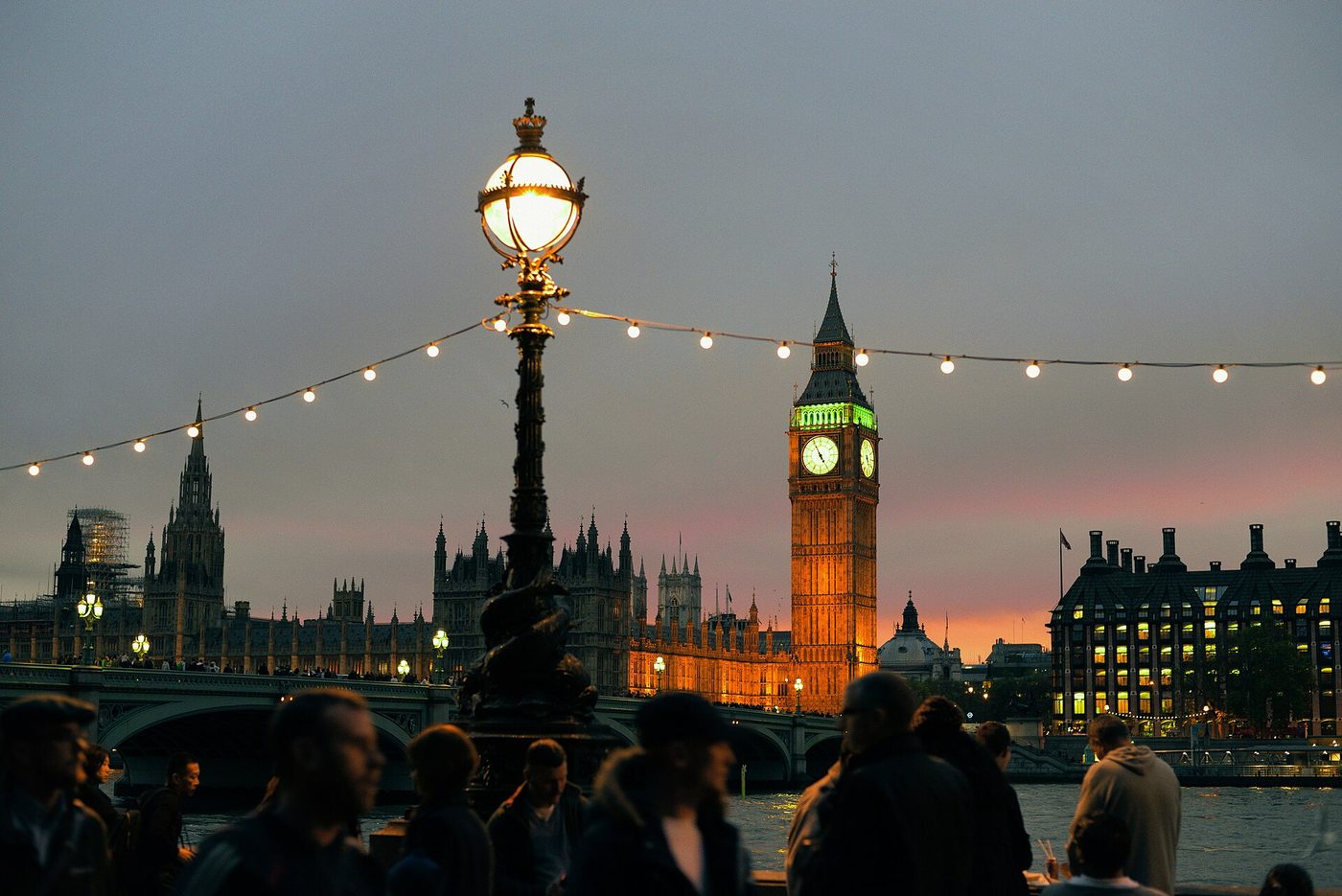 Big Ben and the Houses of Parliament across the Thames at dusk, with strings of lights along the embankment.