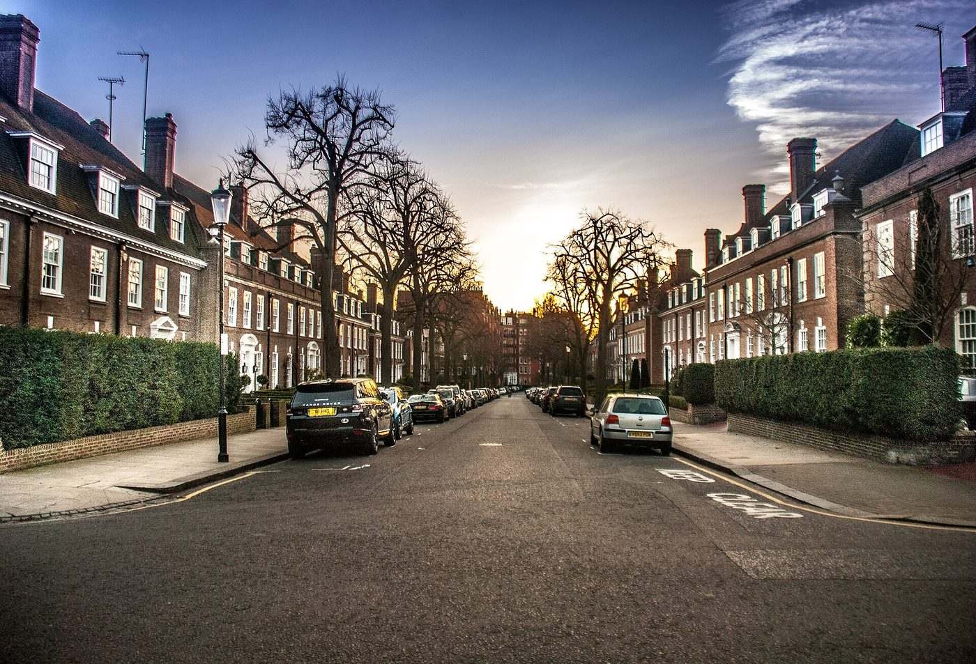 A quiet street of brick terraced houses with tall trees, hedges and parked cars.