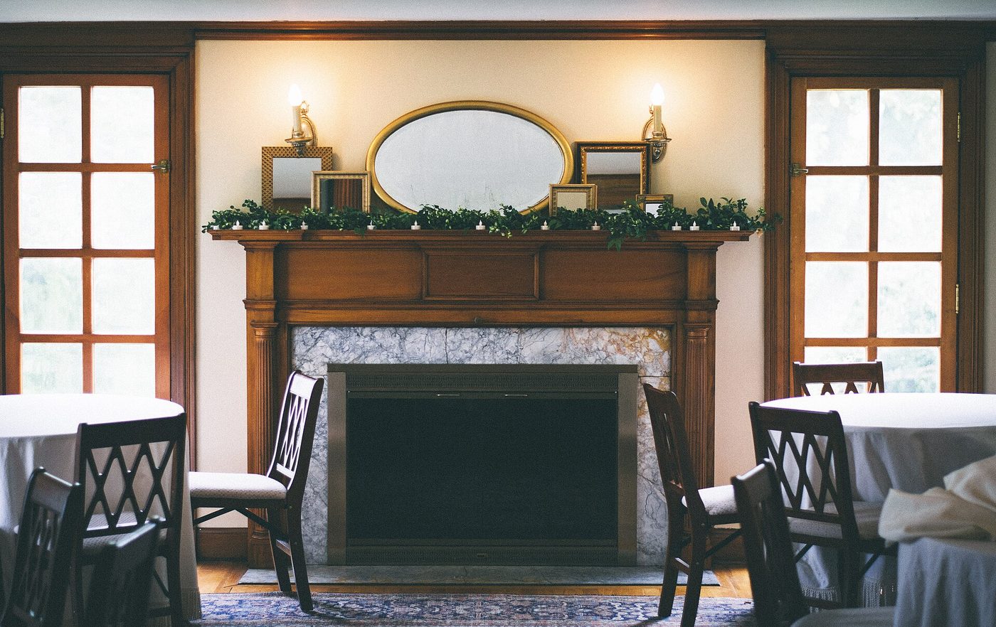 A wooden fireplace with a gilt mirror, wall lights and tall windows on either side, with round tables and chairs in the foreground.