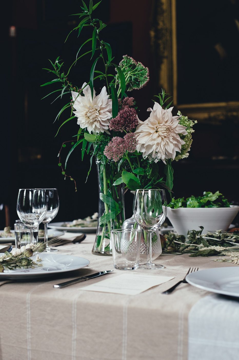 A dinner table set with wine glasses, plates, linen and a vase of dahlias and foliage.