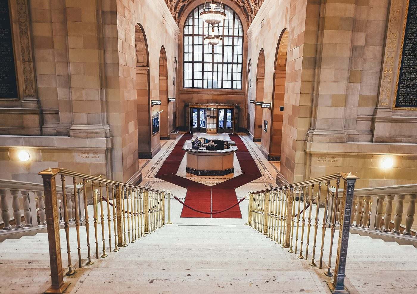 A grand stone staircase with brass banisters and a red carpet leading down to a high, arched hall.